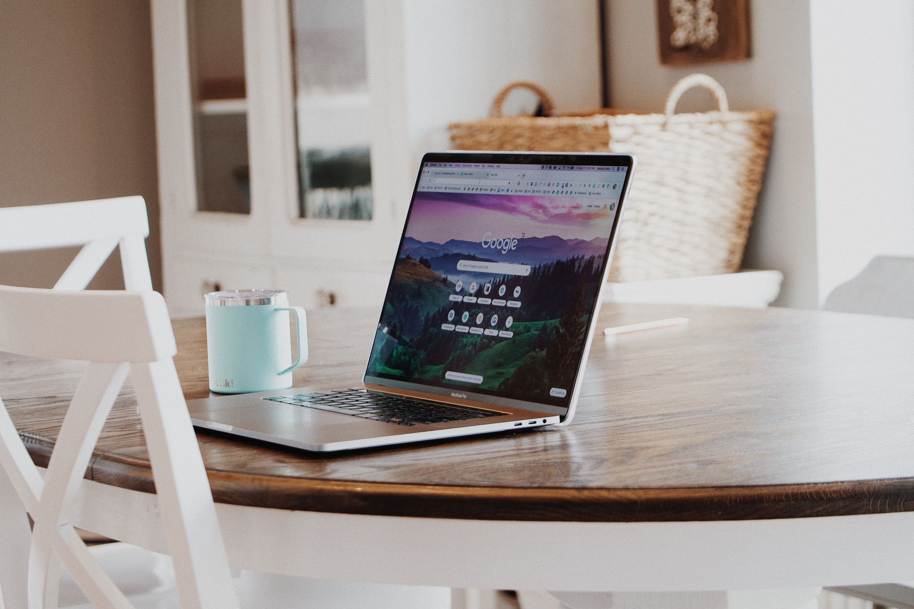 Laptop displaying Google homepage on wooden desk with white chair, mint mug, and woven baskets in background