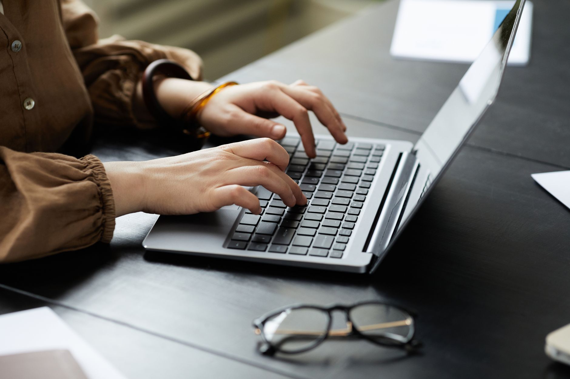 Person typing on a laptop keyboard at a desk with glasses and papers nearby