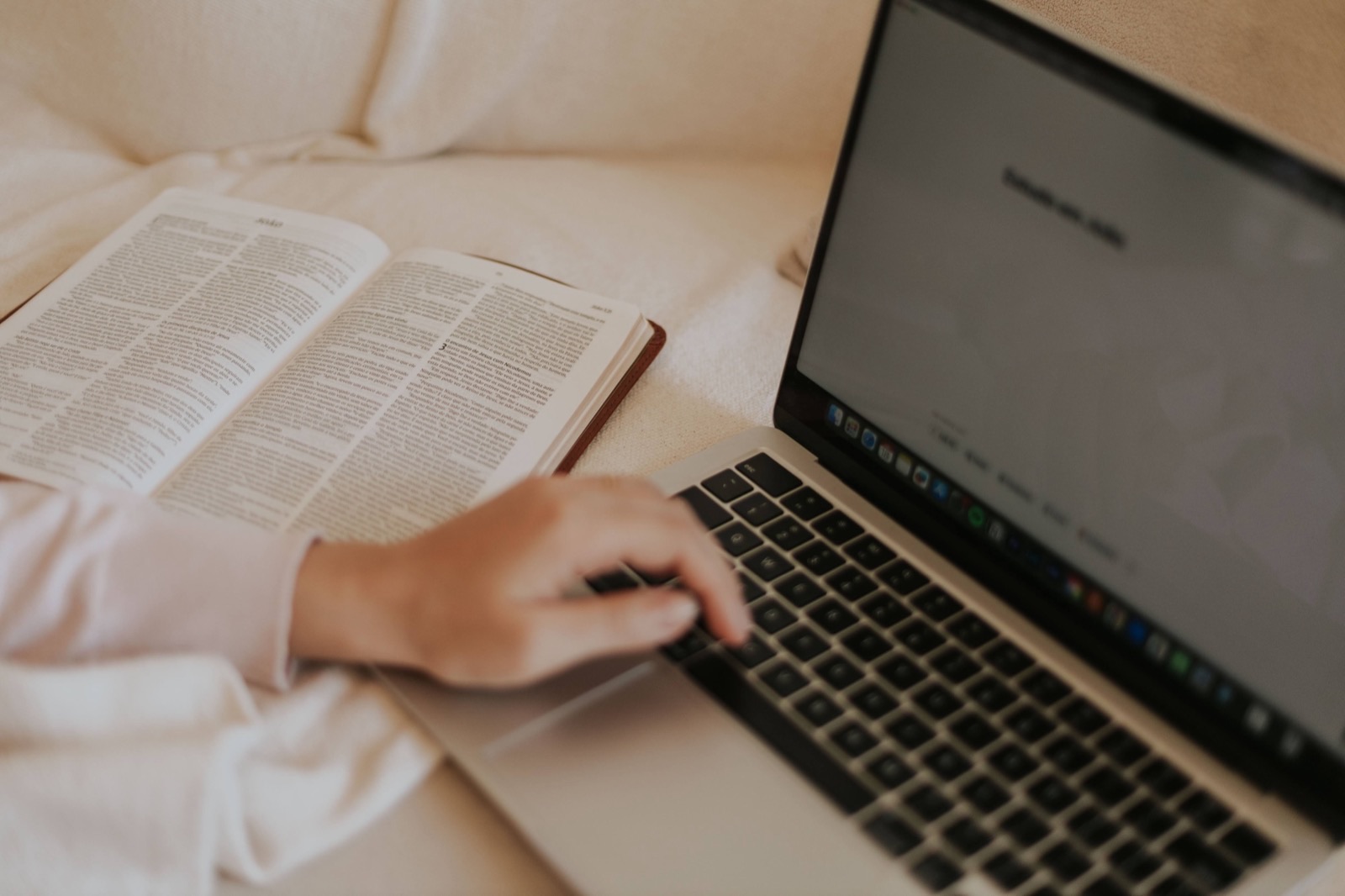 A person in a white sweater reading an open book next to an open laptop on a couch