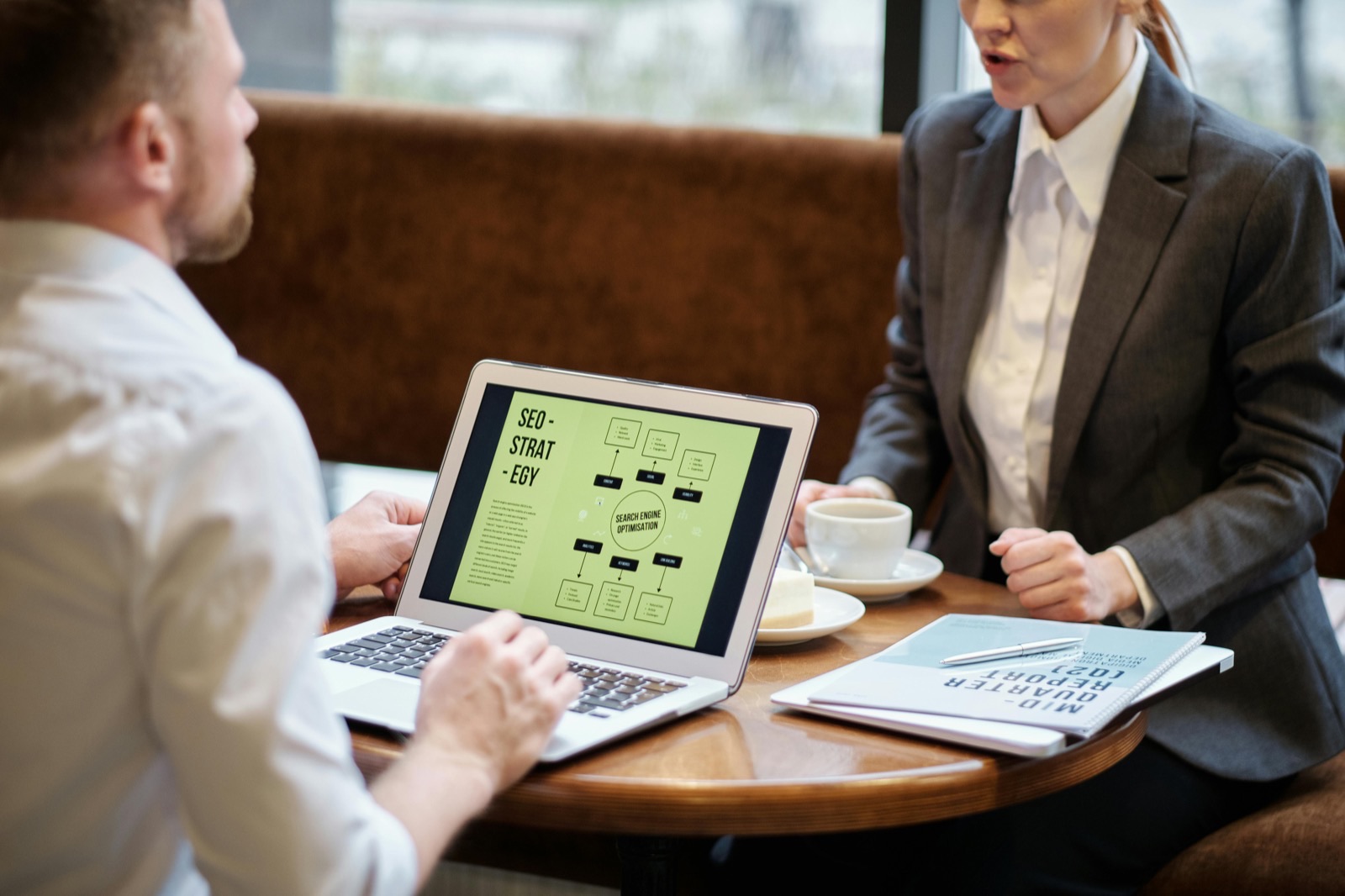 Two professionals discuss SEO strategy while reviewing a flowchart on a laptop during a business meeting at a table with coffee and documents