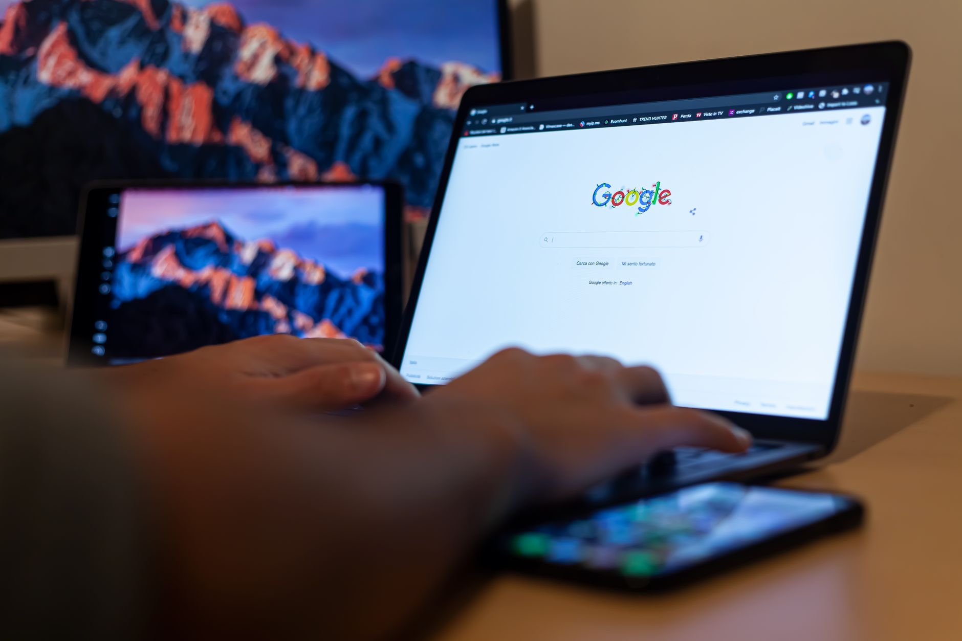 Person using laptop displaying Google homepage with smartphone and tablet nearby on wooden desk