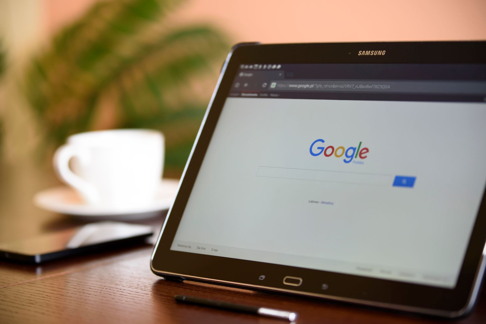Samsung tablet displaying Google search homepage on wooden desk with white coffee cup and potted plant in background
