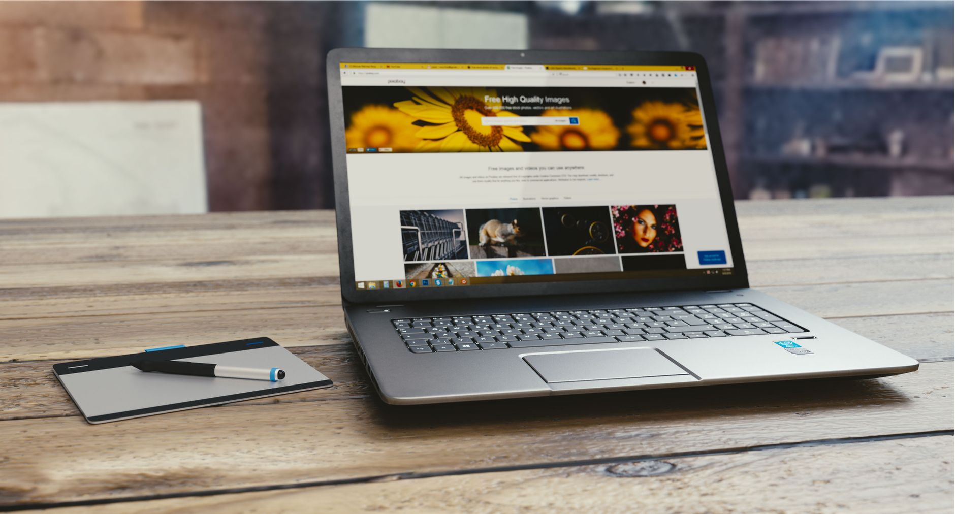 Laptop displaying a free high quality images website with sunflower banner, sitting on wooden desk next to notepad and pen