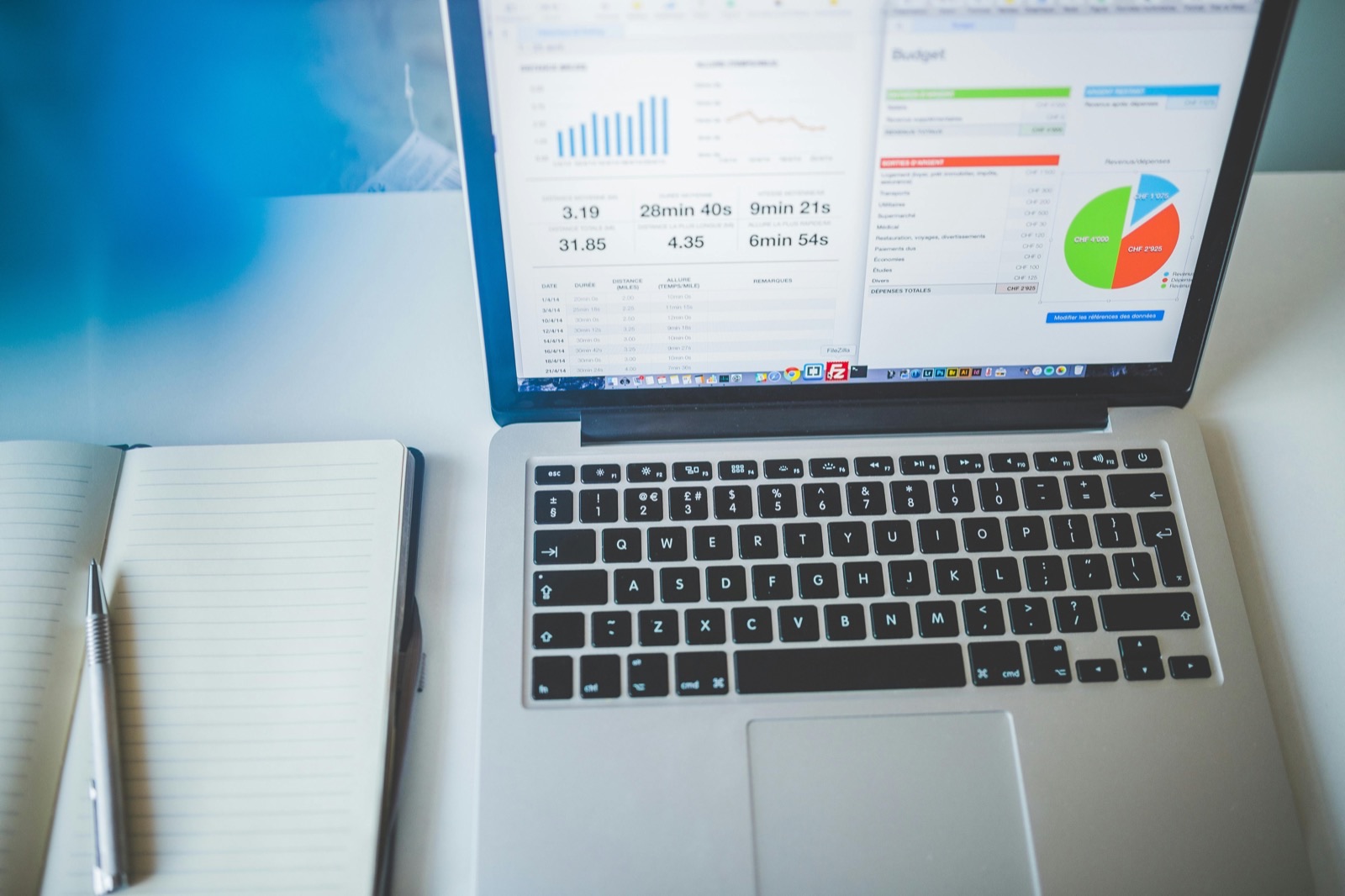 Laptop displaying analytics dashboard with charts and pie graph, next to open notebook and pen on white surface