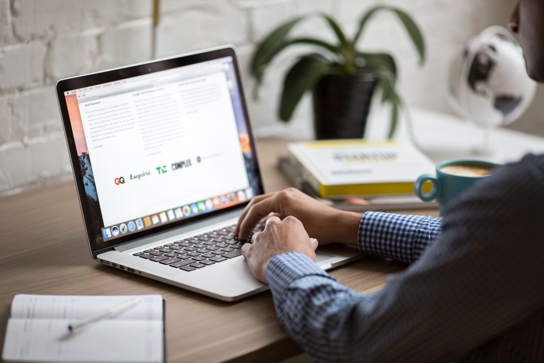 Person in blue checkered shirt working at laptop displaying GQ website at wooden desk with plant, coffee mug, and office supplies