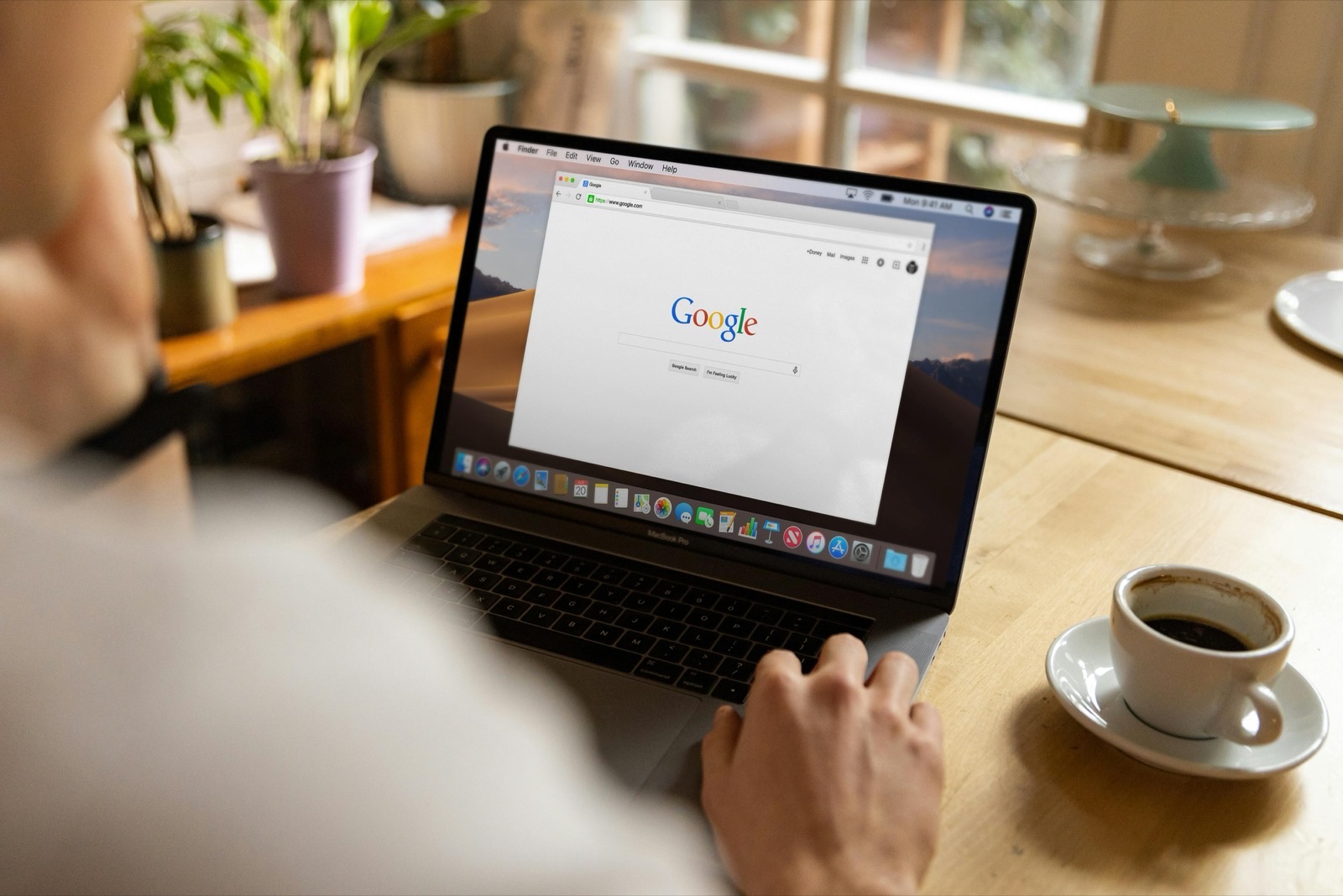 Person using MacBook laptop displaying Google homepage at wooden desk with coffee cup and potted plants