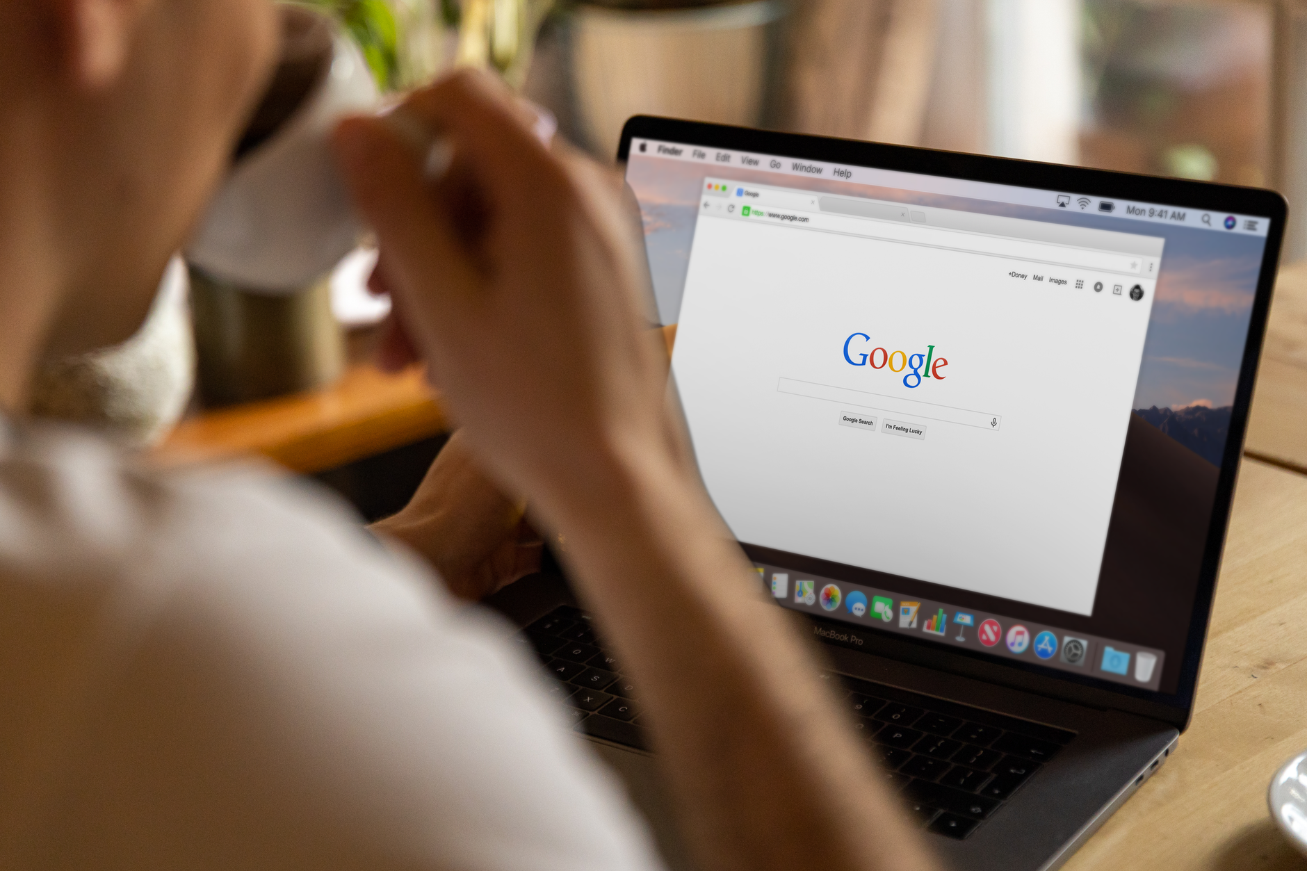 Person sitting at desk with MacBook Pro displaying Google homepage, holding a cup of coffee