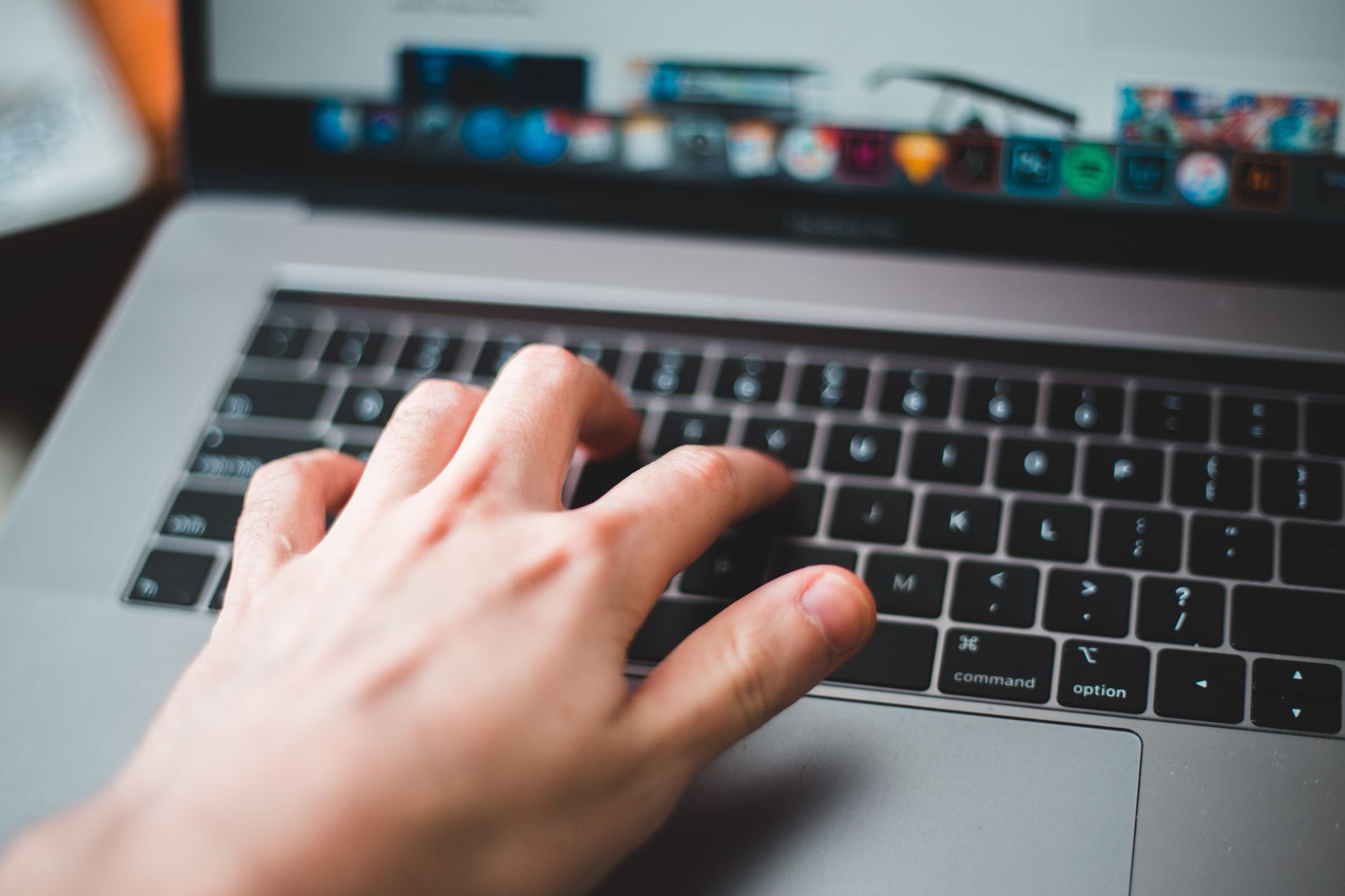 Hand typing on a MacBook keyboard with blurred monitor and desk items in background