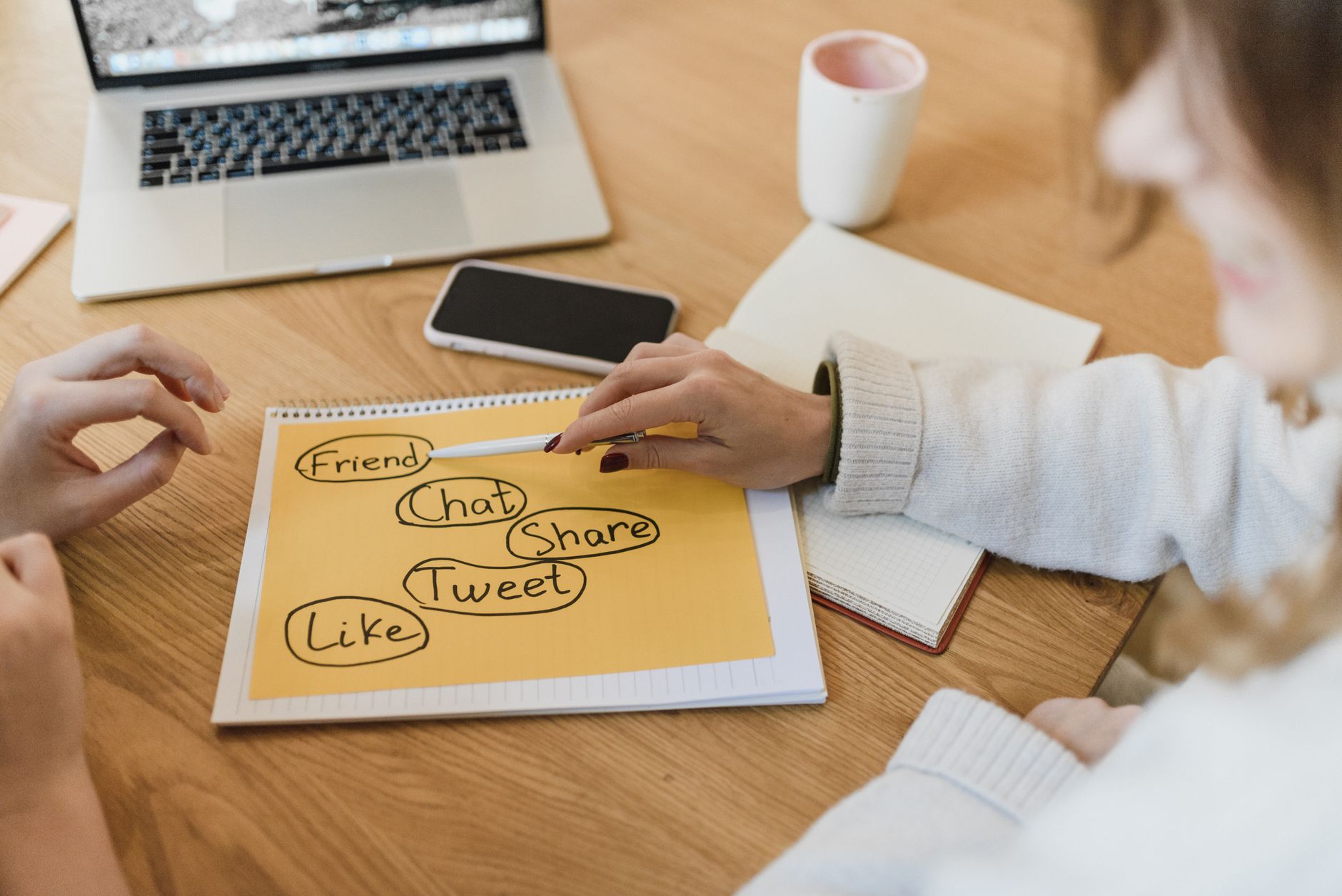 Person writing social media terms on yellow paper at wooden desk with laptop and phone nearby