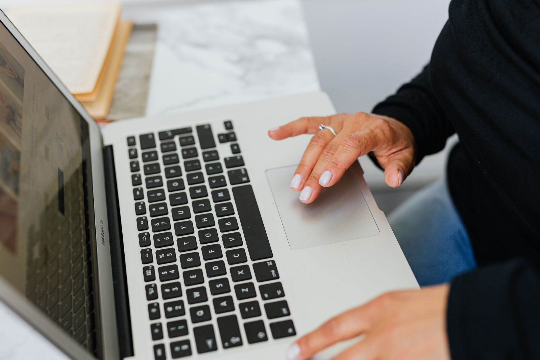 Person using laptop touchpad with manicured nails, wearing black sleeve, with papers and desk visible in background