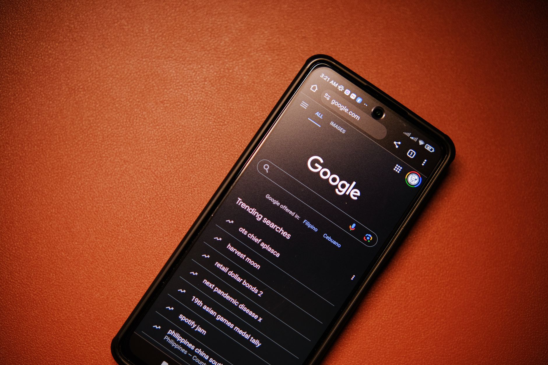 Smartphone displaying Google search homepage in dark mode with trending searches listed below the Google logo on an orange textured background