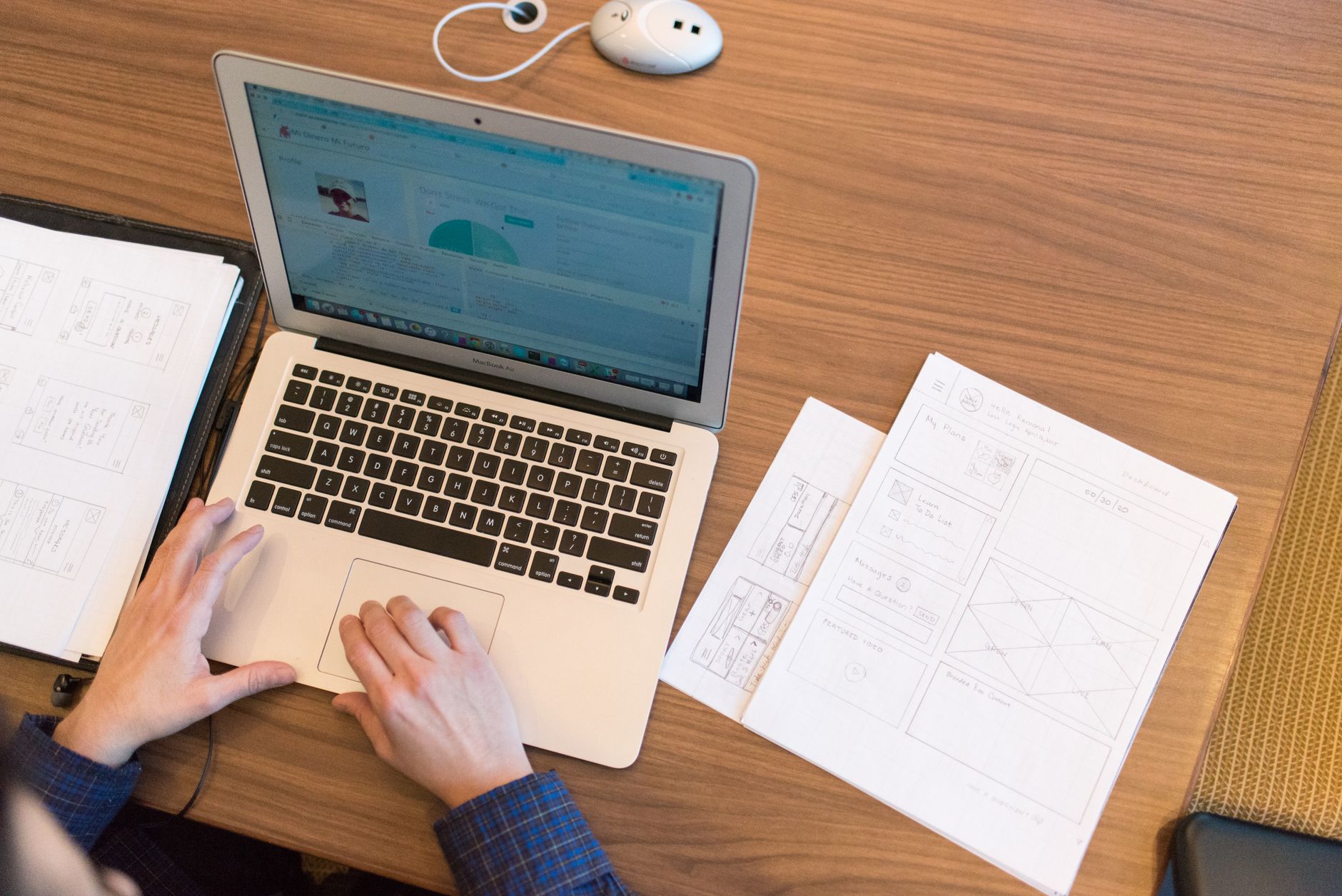 Person using MacBook at wooden desk with wireframe sketches and mouse nearby