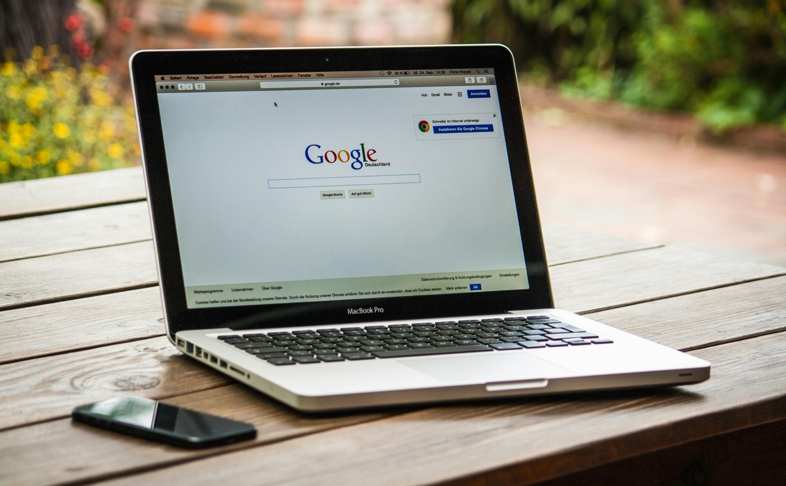 MacBook Pro on wooden table displaying Google Deutschland homepage with smartphone beside it and garden in background