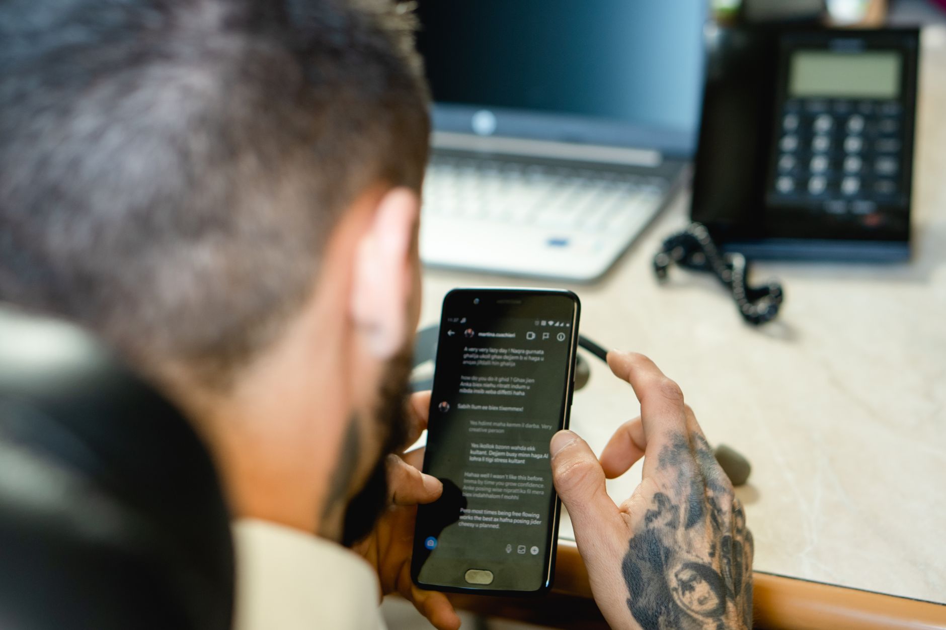 Person with tattoo holding smartphone displaying text, with laptop and desk phone blurred in background