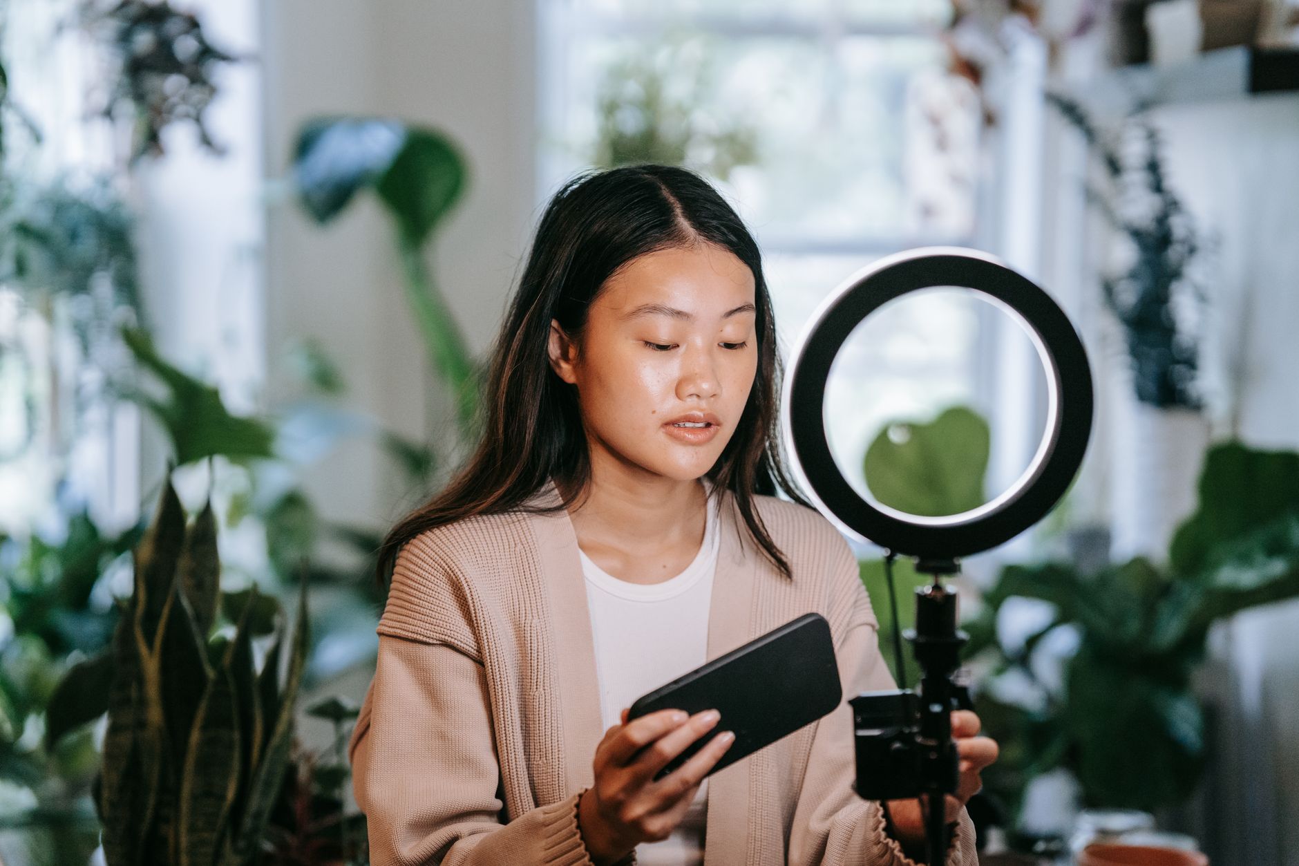 Young woman holding a smartphone and ring light surrounded by potted plants in a bright room