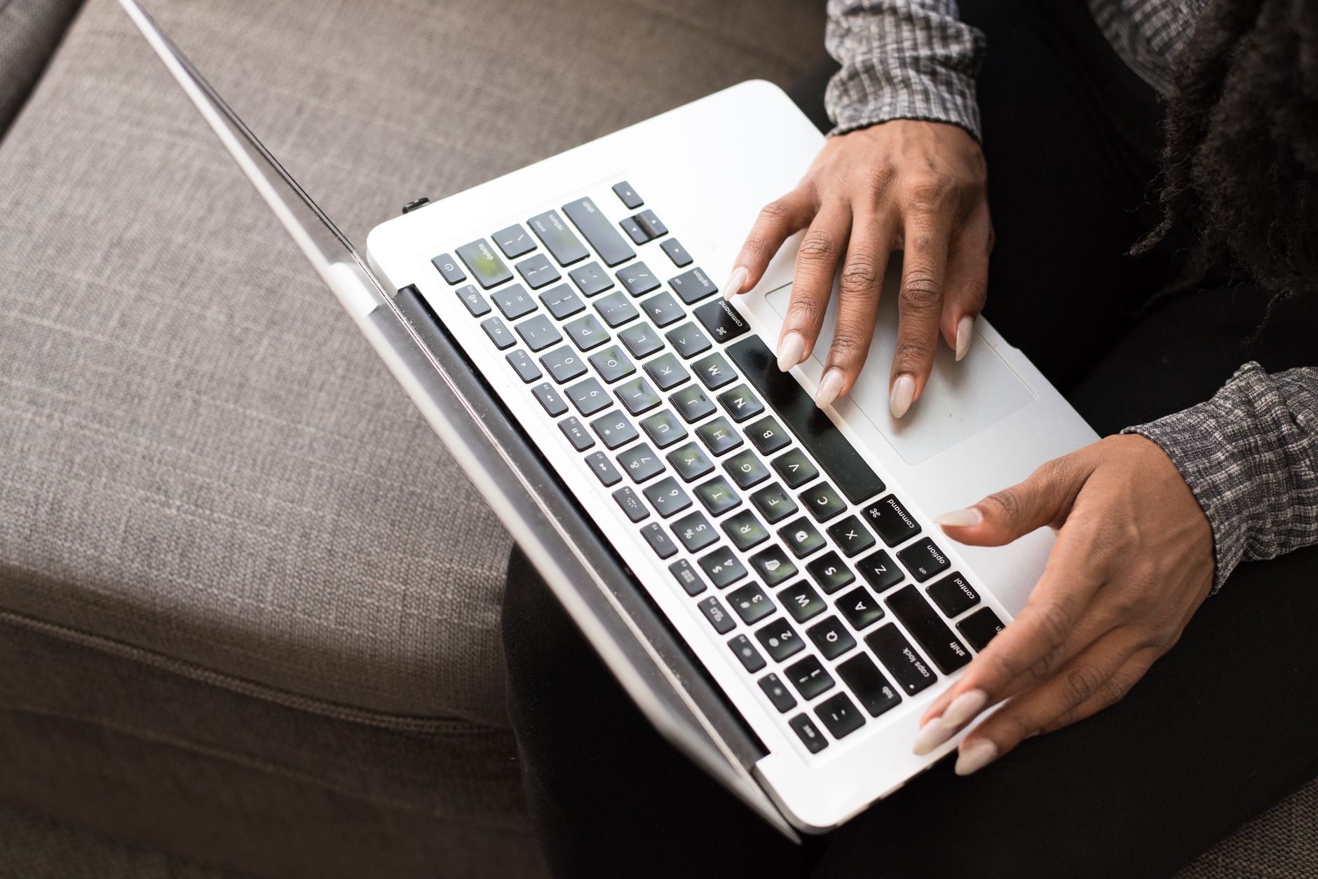 Person in checkered shirt using white laptop while sitting on gray couch