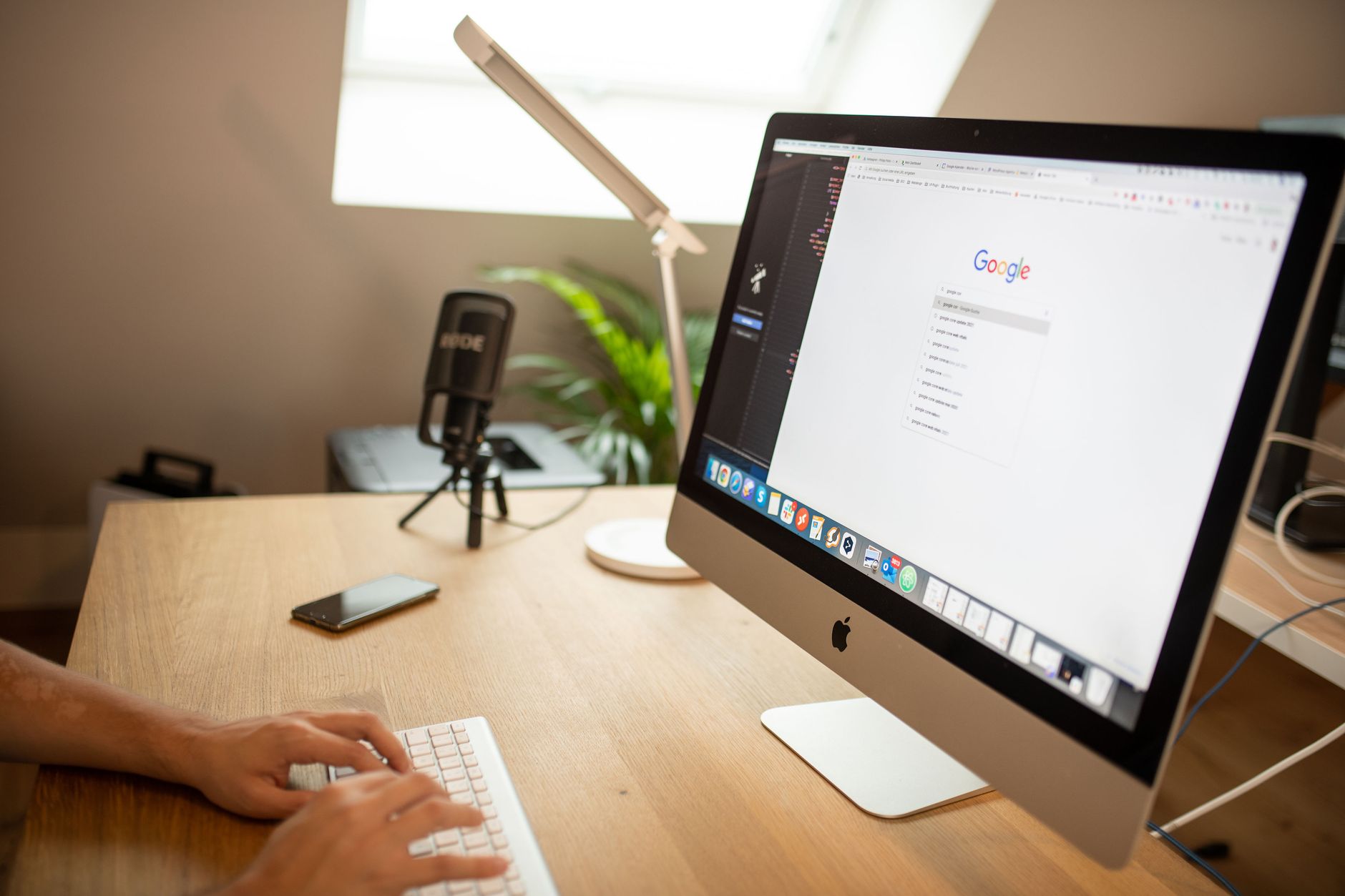 Person using keyboard at wooden desk with iMac displaying Google homepage, microphone, lamp, and potted plant visible