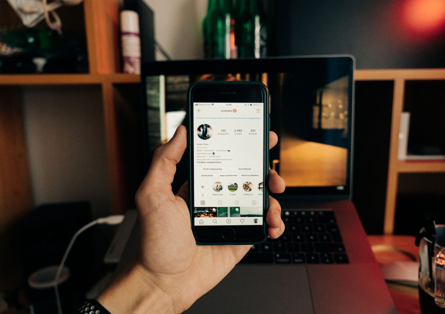 Hand holding iPhone displaying an Instagram profile with 132 followers, 5,465 following, and 331 posts, in an office setting