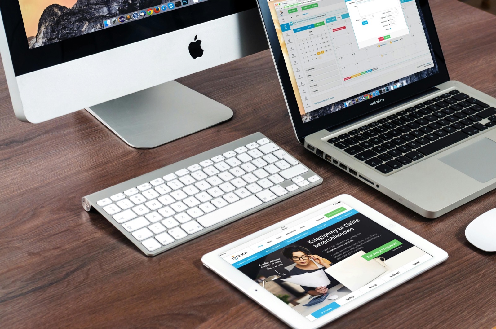 An iMac, a MacBook laptop, a wireless keyboard and an iPad displaying a website on a wooden desk