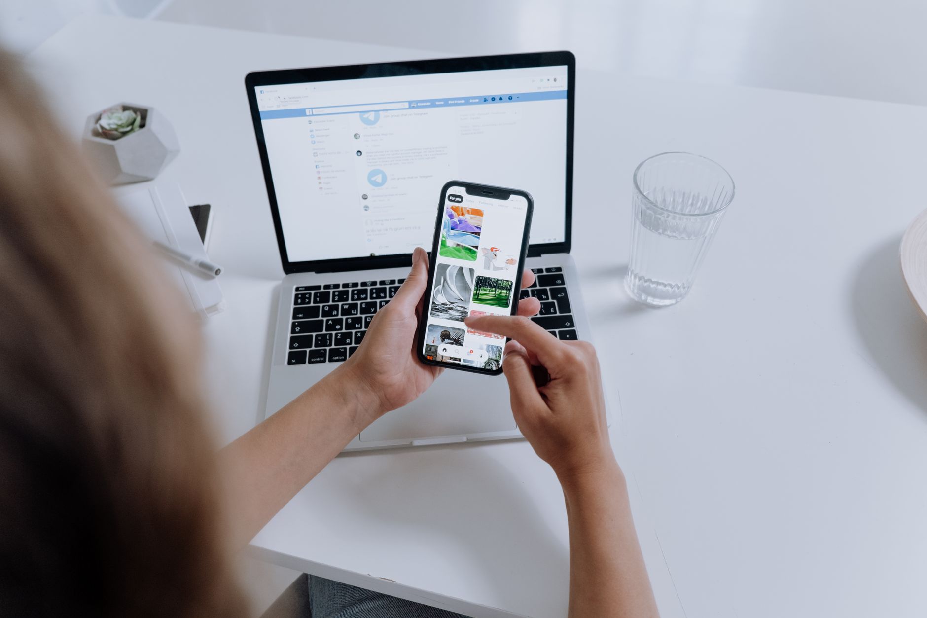 Person holding smartphone displaying social media app above laptop with open browser on white desk