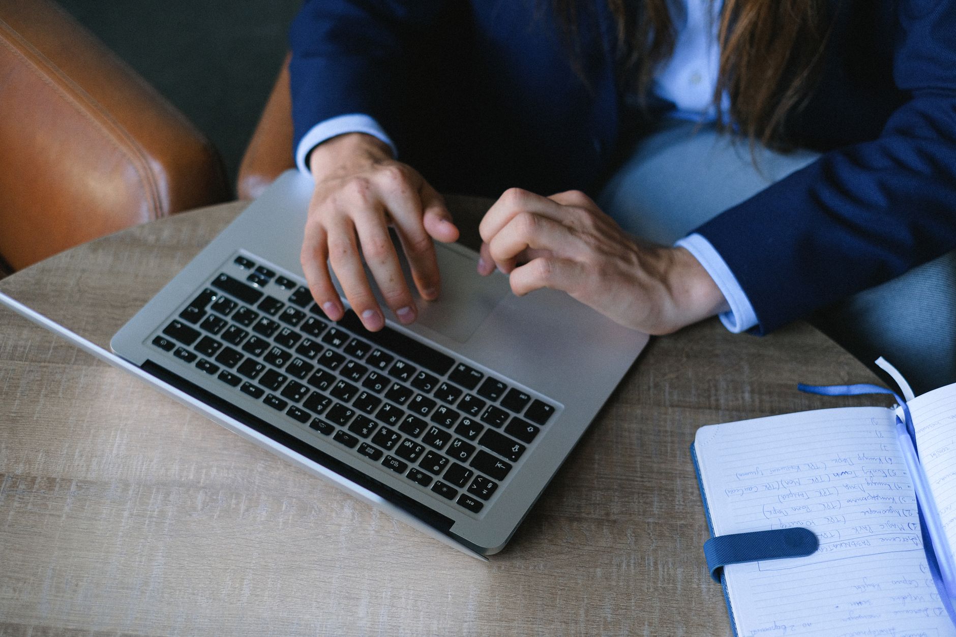 Person in blue business suit typing on laptop at desk with notebook and pen nearby