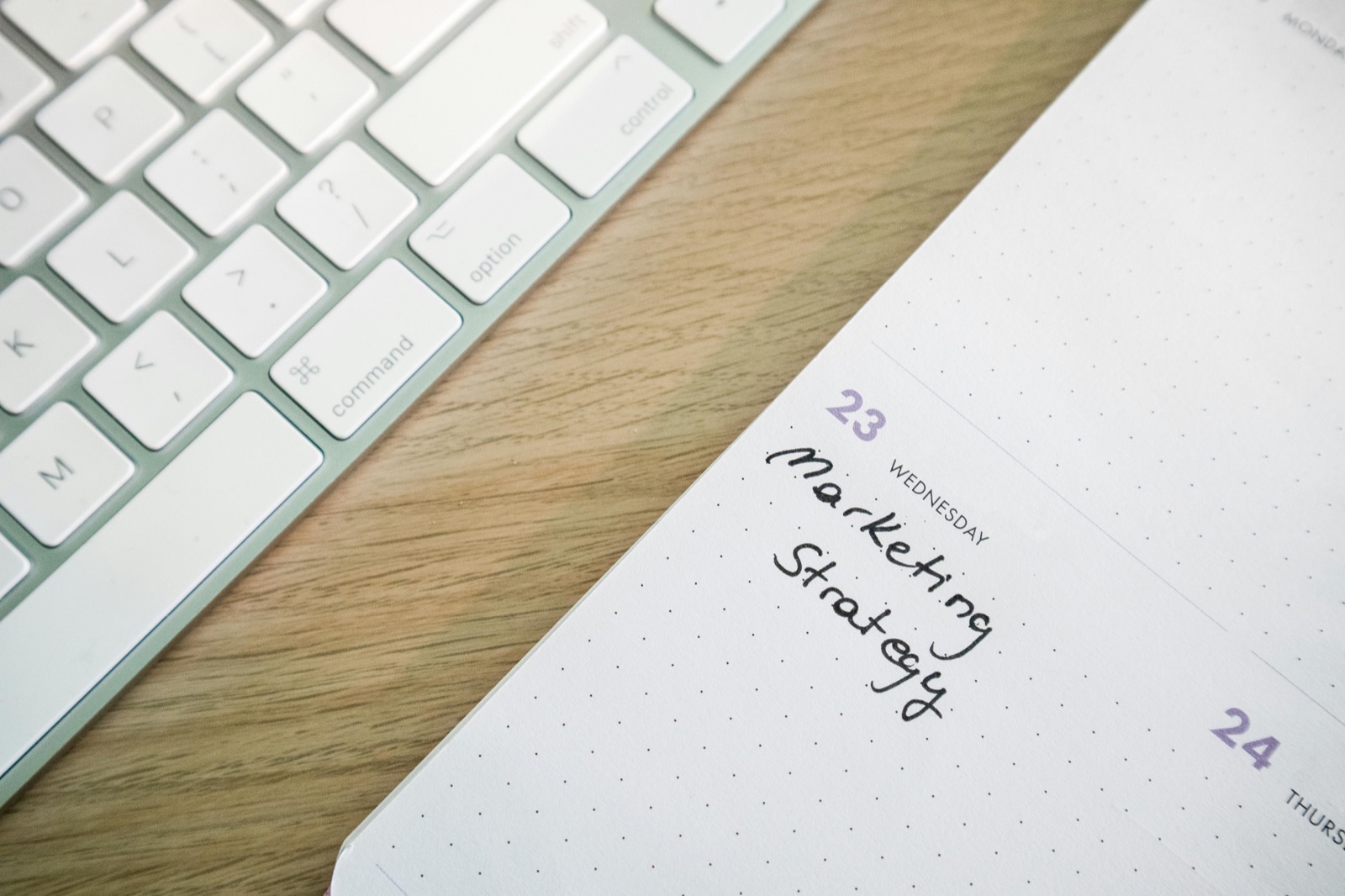 A white keyboard and an open planner showing Wednesday the 23rd with handwritten text reading "marketing strategy