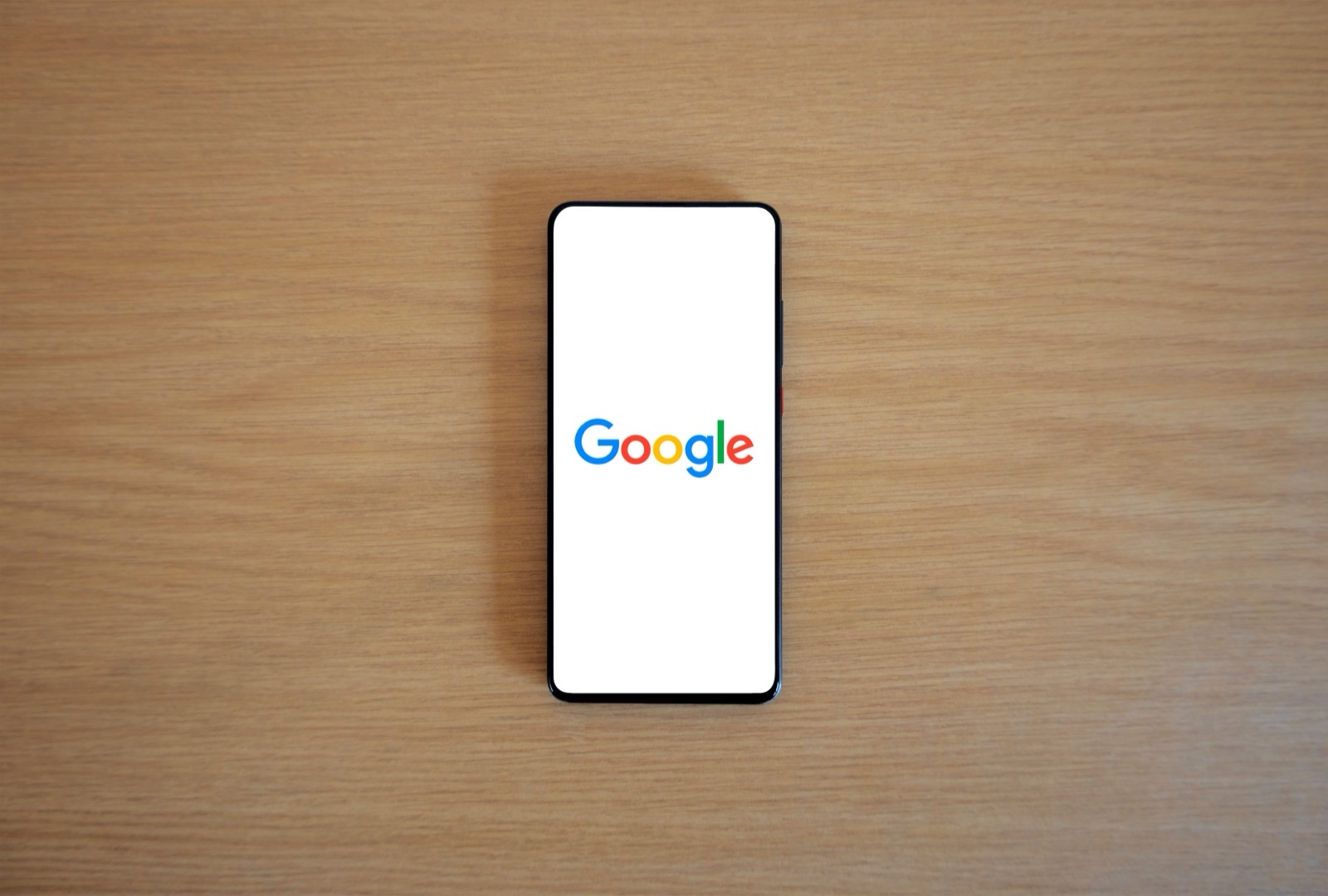 Smartphone with black frame displaying Google logo on white screen, centered on wooden surface