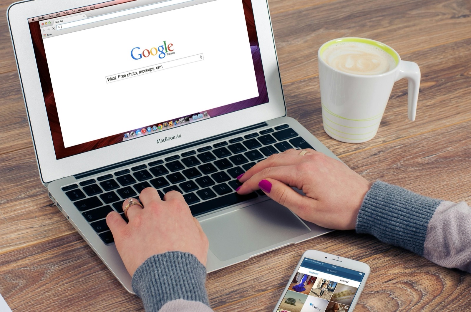 Person using MacBook Air displaying Google homepage, with coffee mug and smartphone on wooden desk