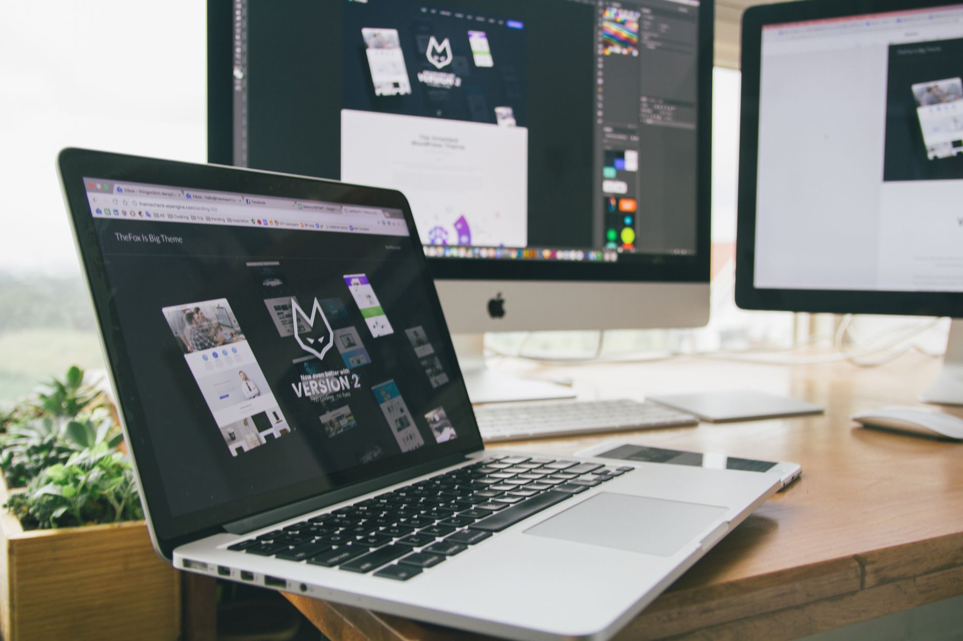 Designer workspace with MacBook laptop and iMac displaying website mockups and design projects with dark theme interface