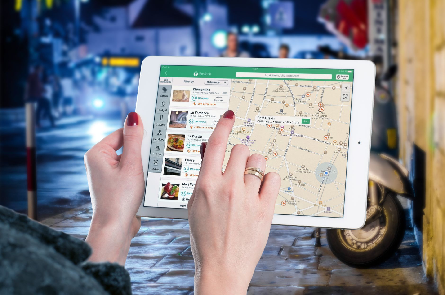 Person holding white tablet displaying TheFork restaurant app with map of Paris and restaurant listings on street background