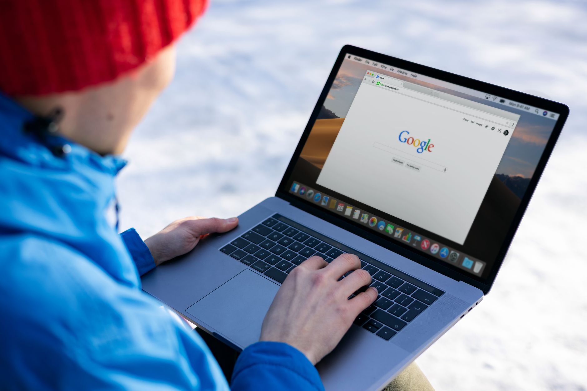 Person in blue jacket and red hat using laptop displaying Google homepage outdoors in snowy setting