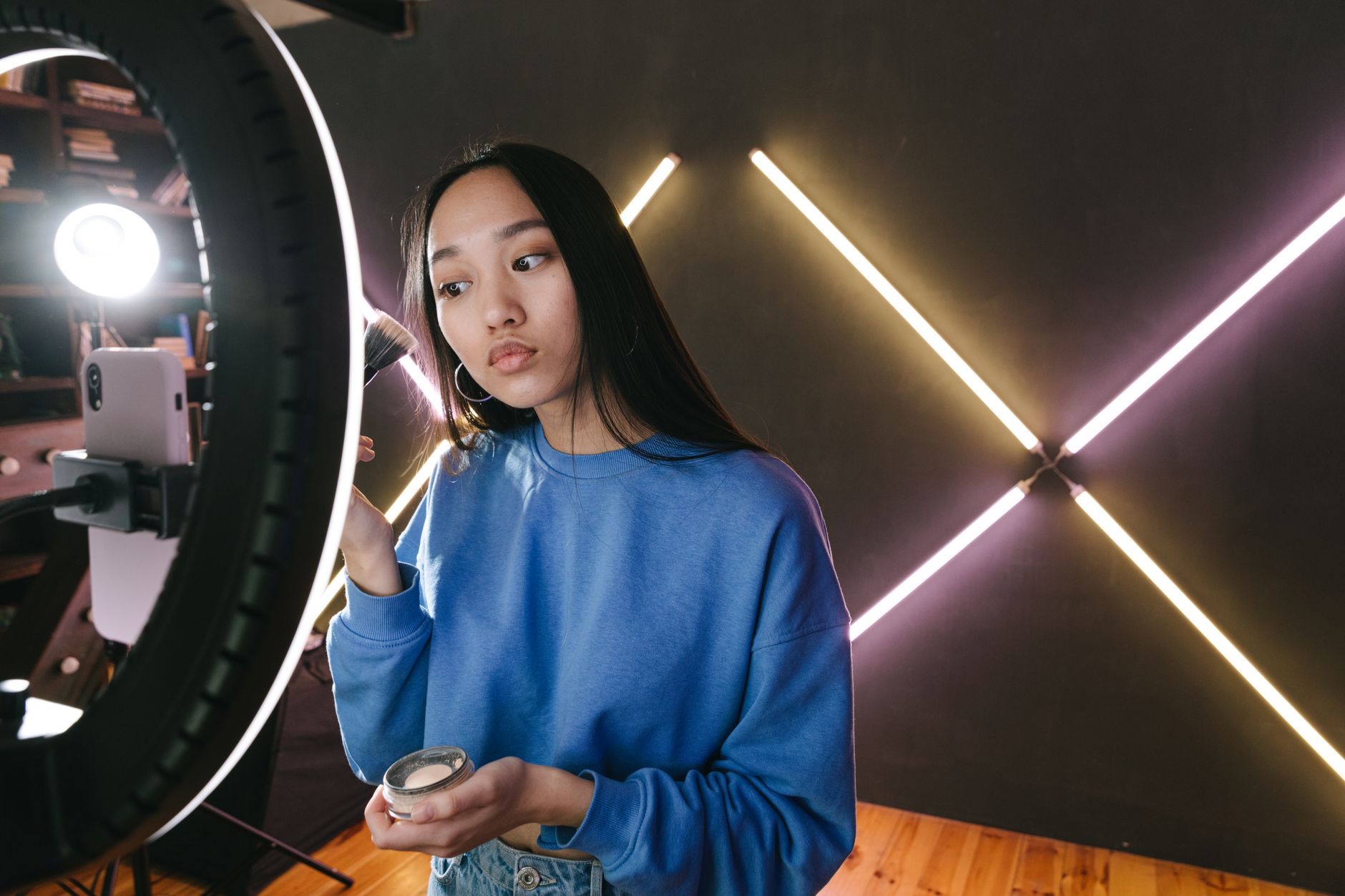 Woman in blue sweatshirt holding a ring light while recording with a smartphone in front of neon geometric lighting