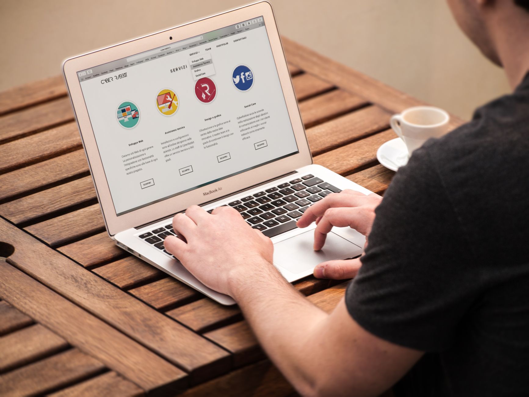 Person using MacBook Air laptop displaying website with colorful service icons on wooden table with coffee cup