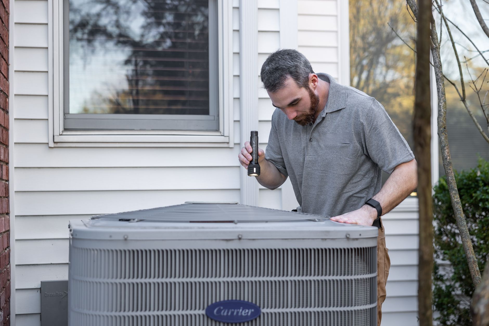 An HVAC technician using a flashlight to inspect inside a residential outdoor condenser unit